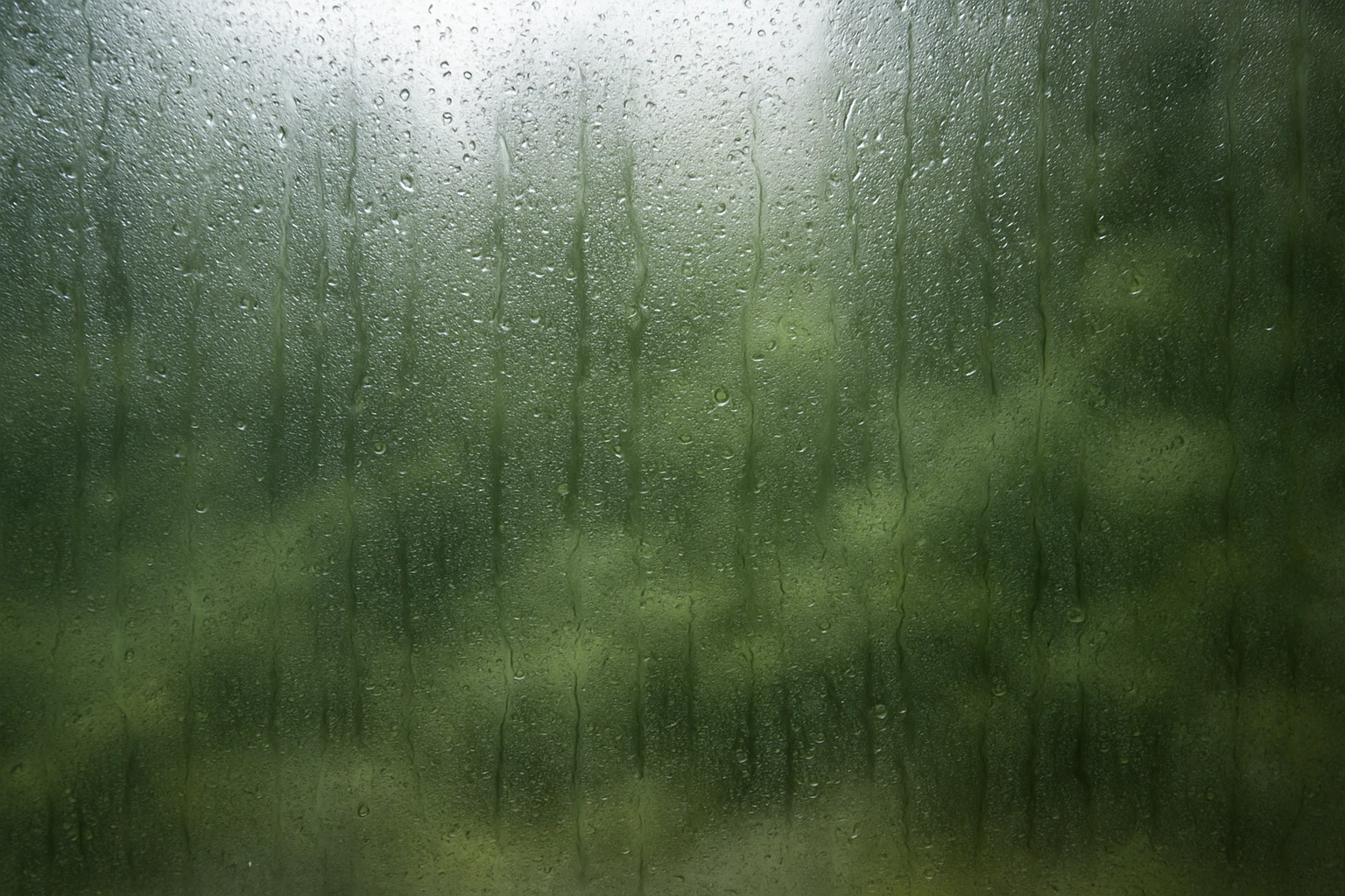 Egy esős ablakon keresztül látható zöld táj, vízcseppekkel borítva.
